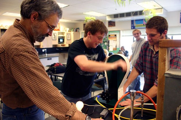 South Whidbey High School physics teacher Greg Ballog shows Kellen Field and Patrick Parnell how the hand-crank generator powers a small light bulb. The equipment