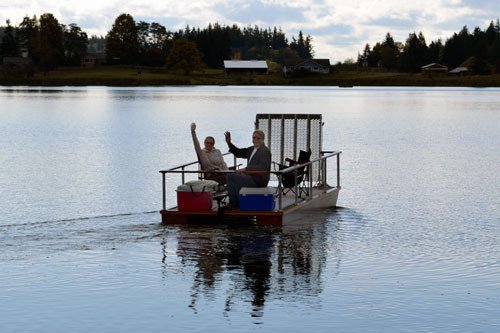 Mike and Anne Mayes wave as they ride the ‘Accessible’ across the water of Lone Lake to the boat’s host property.