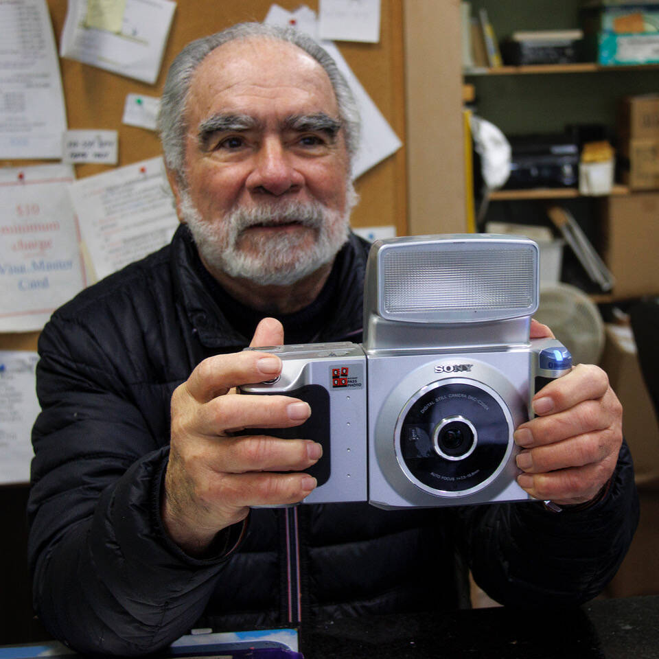 Photo by David Welton
George Guzman, seen here with his camera, has taken countless passport photos over the years. He is retiring from his downtown Langley business, GHL Enterprises.