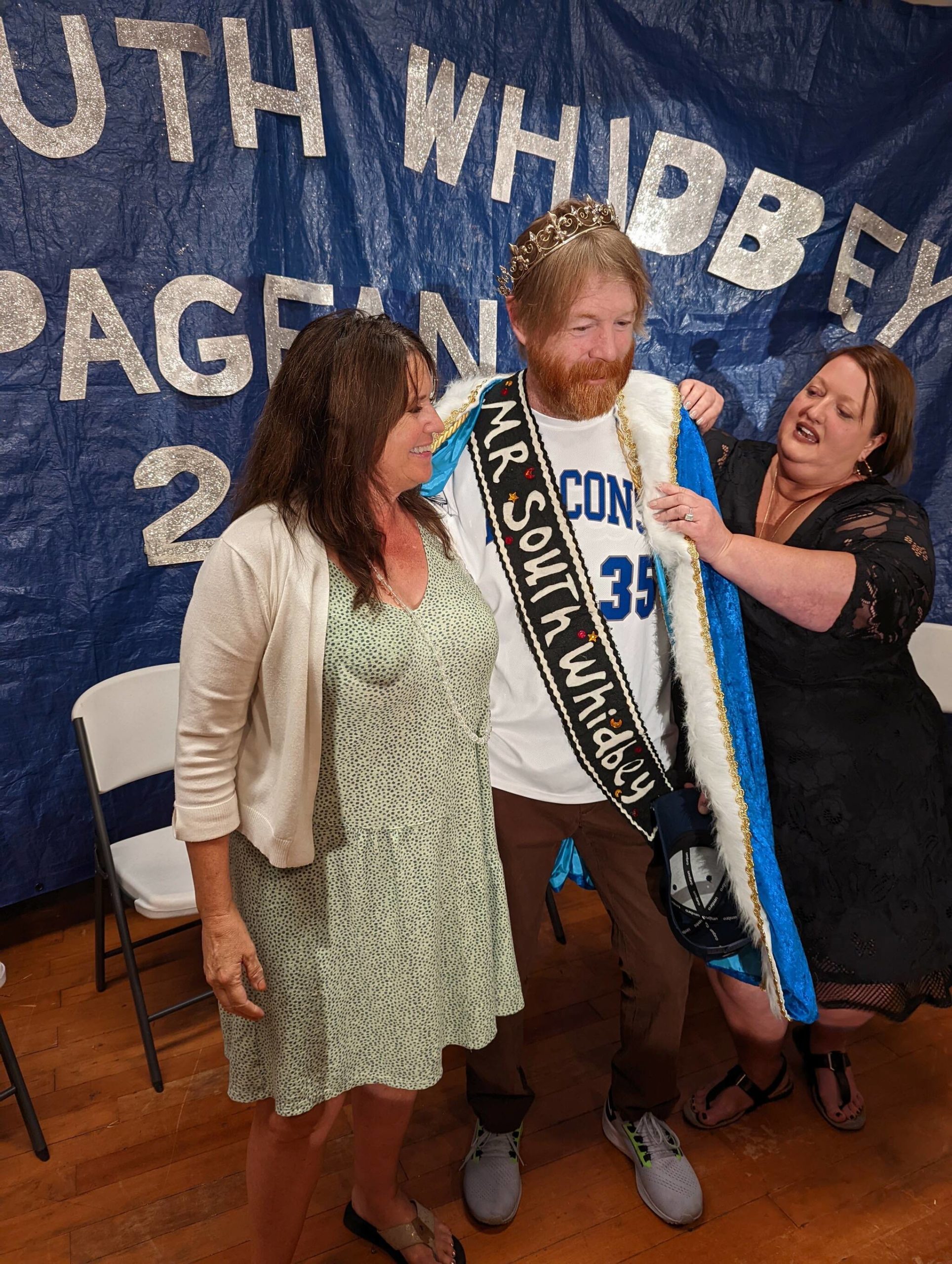 Photo by Sue Frause
Erik Stine, standing in for John LaVassar, was crowned Mr. South Whidbey this past weekend. Kristi Price, the board president for Friends of Friends Medical Support Fund, helped Stine into the Mr. South Whidbey robes while Gail, LaVassars wife, stood nearby.