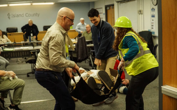 Island County Planning Manager Michael Jones (left), Health Director Shawn Morris (back) and Board Clerk Jennifer Roll (right) carry an actor during an earthquake demonstration at the commissioner's meeting on Wednesday. (Photo by Sam Fletcher)