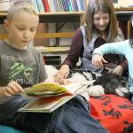 Evan Thompson / The Record &mdash; Zane Floyd, 8, reads a book to Ruby, a three-year-old parti poodle and therapy dog Reading with Rover. Nicholle Sargeant, 7, and handler LouAnn Hepp gently brushes Ruby&rsquo;s fur.