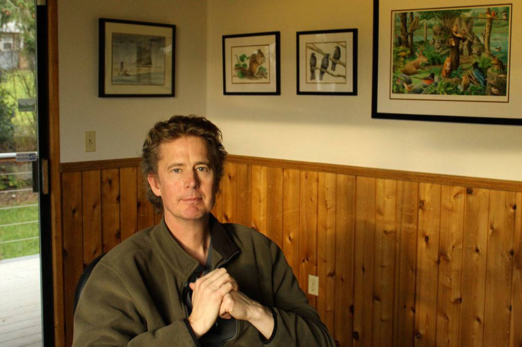 Craig Johnson poses beneath a wall of his artwork in the couples home. (Photo by Patricia Guthrie/Whidbey News Group)