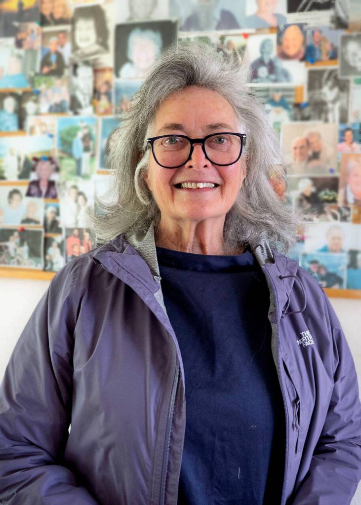 Photo by David Welton
Retiring Executive Director Ann Cutcher stands before a wall of photos of guests who have stayed at Enso House.